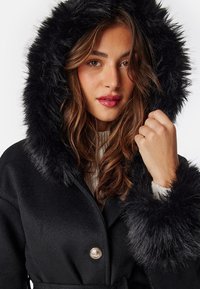 Black coat with a faux fur hood and cuffs. It features a wrap design and is adorned with a decorative button at the collar.