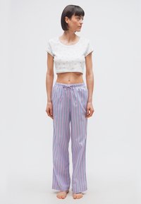 Femme aux cheveux courts et foncés, portant un crop top blanc et un pantalon de pyjama rayé violet clair, debout pieds nus et regardant sur le côté.