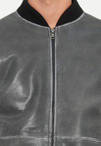 Chaqueta bomber de cuero gris con una textura suave, cuello negro acanalado y cremallera frontal. Presenta una costura mínima y sin bolsillos visibles.