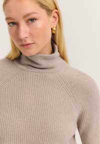 Beige Rollkragenpullover mit strukturiertem Rippdesign, ausgestattet mit Raglanärmeln und einer figurbetonten Passform. Goldfarbene Ohrstecker ergänzen den Look.