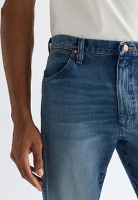 Nærbilde av en person iført blå denim jeans og en hvit skjorte, med fokus på midjen, lommen og venstre arm.