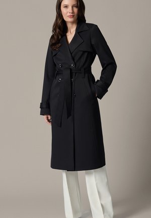 Kobieta w długim czarnym dwurzędowym płaszczu trenczowym z paskiem, zestawionym z białymi spodniami o szerokich nogawkach, stoi na jednolitym tle.