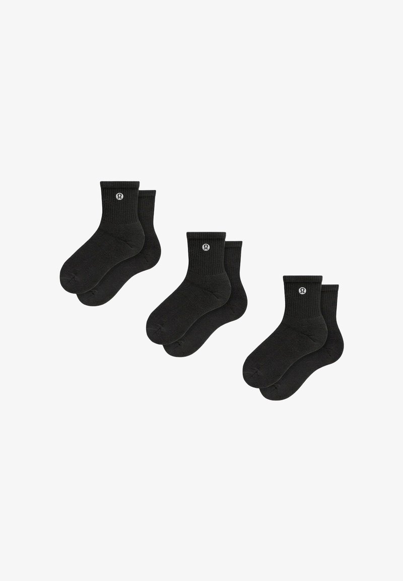 Trois paires de chaussettes noires jusqu'au milieu du mollet avec des poignets côtelés et un petit logo circulaire blanc sur chaque poignet, présentées sur un fond blanc.