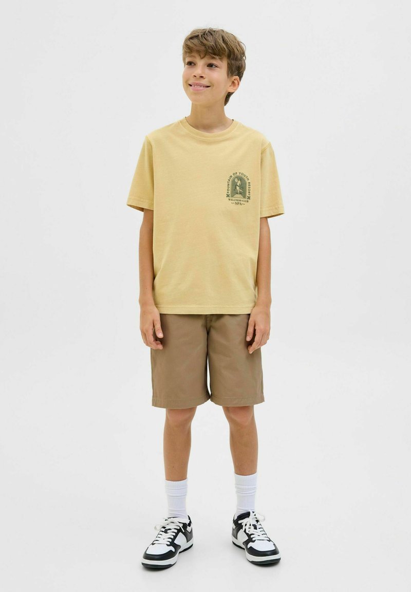 T-shirt en coton jaune avec un motif vert, associée à un short beige. Des chaussettes blanches et des baskets noir et blanc complètent la tenue.