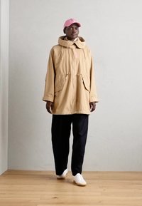 Beige utendørsjakke med hette, to sidelommer og snor. Brukt med svarte bukser og hvite sneakers. Akkompagnert av en rosa caps.
