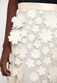 Main à la peau foncée à côté d'une jupe en tissu transparent ornée d'appliques florales blanches 3D superposées de différentes tailles.