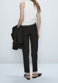 Czarne dopasowane spodnie o prostym kroju, zestawione z dopasowaną białą koszulą bez rękawów i czarnym żakietem zarzuconym na jeden ramię. Czarne płaskie buty.