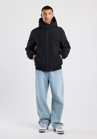 Schwarz isolierte Jacke mit Kapuze, Frontreißverschluss und elastischen Bündchen, kombiniert mit hellblauen weit geschnittenen Jeans und weißen Sneakers.