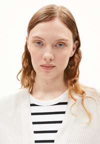 White knitted cardigan over a black and white striped shirt. Model has wavy, medium-length hair and is facing forward. Neutral background.