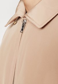 Ljust beige zip-jacka med spetsig krage, utrustad med en silverdragkedja och en slät, blank yta.