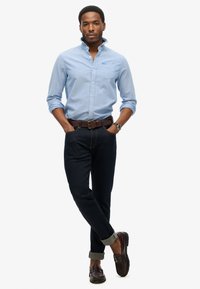 Lichtblauw overhemd met knopen en een borstzak, donkerdenim jeans opgerold bij de enkels, en bruine instappers met een gestructureerde afwerking.