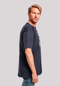 Navy blue cotton t-shirt with short sleeves and a loose fit, featuring white text graphic on the front. Model stands sideways.