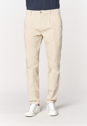 Pantaloni beige slim fit indossati con sneakers bianche e camicia blu navy, mostrati dalla vita ai piedi su uno sfondo uniforme.