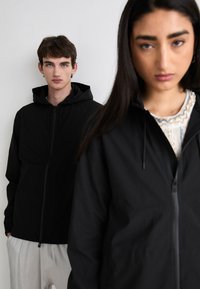 Jachetă cu fermoar neagră, cu glugă și șnururi, textură netedă; modelul din față poartă un top cu model, ambele pe un fundal alb simplu.