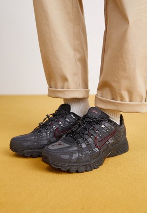 Osoba nosząca beżowe spodnie i białe skarpetki z czarnymi butami Nike z czerwonymi logo w kształcie łyżwy na podłodze w kolorze musztardowym.