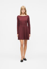 Νεαρή γυναίκα που στέκεται στραμμένη προς τα εμπρός, φορώντας ένα μακρυμάνικο φόρεμα μήκους μέχρι τα γόνατα σε μπορντό χρώμα και μαύρα μπαλαρίνα παπούτσια, πάνω σε λευκό φόντο.