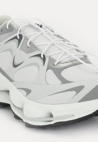 Pantofii sport albi cu accente gri, partea superioară din plasă și detalii texturate. Au șireturi plate și o talpă căptușită pentru susținere.