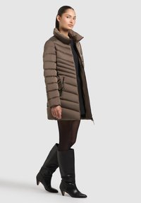 Maslinasti puffer kaput s visokim ovratnikom, kviltanim dizajnom, zatvorenim džepovima i elegantnim krojem, uparen s crnim kožnim čizmama do koljena.