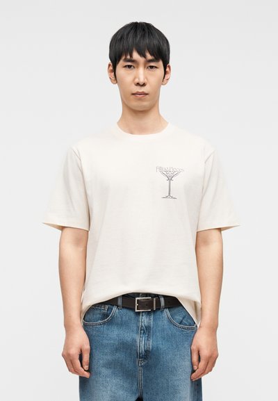 Μπεζ βαμβακερό μπλουζάκι με κοντά μανίκια, με μαύρο γραφικό σχέδιο ποτηριού κοκτέιλ στο στήθος. Φορεμένο με μπλε τζιν παντελόνι.