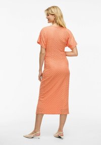 Robe midi texturée pêche à manches courtes et silhouette ajustée ; présente un subtil motif géométrique. Portée avec des chaussures à enfiler beiges.