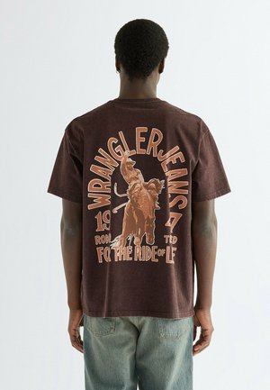 Persona che indossa una t-shirt Wrangler Jeans marrone con una grafica di un cavaliere del rodeo e il testo "For the ride of life" sulla schiena, abbinata a jeans azzurri chiari.