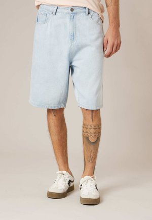 Homme portant un short en denim bleu clair, des baskets blanches avec des accents noirs, et des tatouages géométriques visibles sur les deux jambes.