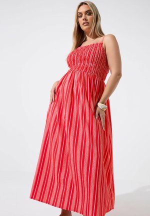 Femme portant une longue robe sans manches rayée verticalement rouge et blanche avec un corsage ajusté et une jupe fluide, debout devant un fond uni.