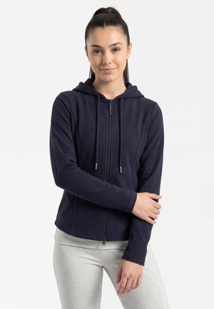 Jonge vrouw met donker haar in een staart draagt een marineblauwe zip-up hoodie en lichtgrijze broek, staat met één arm over haar lichaam gekruist.