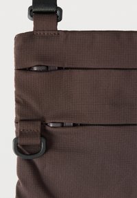 Hnedá látková taška s dvoma vodorovnými zipsovými vreckami, štruktúrovaným mriežkovým vzorom a čiernym kovovým D-krúžkom na popruhu.