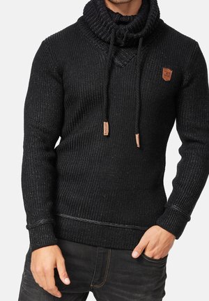 Mężczyzna ubrany w czarny prążkowany sweter z wysokim golfem i sznurkami, w zestawieniu z czarnymi dżinsami, pozujący z jedną ręką w kieszeni.