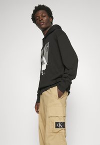 Felpa nera con cappuccio e stampa grafica; indossata con pantaloni cargo beige caratterizzati da un patch con logo sulla coscia; vestibilità comoda, materiale in cotone.