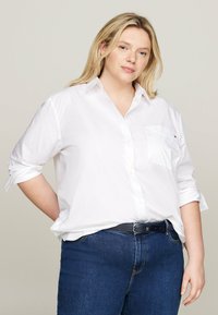 Biela košela s krátkymi rukávmi a náprsním vreckom, v kombinácii s tmavomodrými rifľami a čiernym opaskom. Jednoduchý a klasický dizajn.