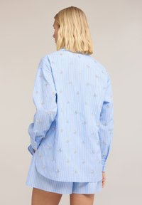 Persona rubia de espaldas lleva camisa azul claro con rayas y pantalones cortos a juego con pequeños bordados florales blancos en la camisa.