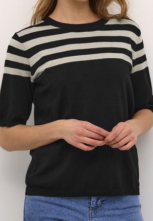 Femme portant un pull noir à manches courtes avec trois bandes horizontales beiges sur les épaules et le haut de la poitrine, associé à un jean bleu.