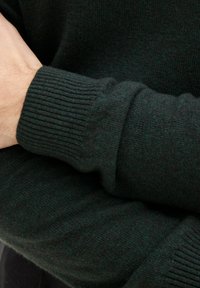 Dunkelgrüner Pullover mit gerippten Bündchen, aus einem weichen Strickmaterial gefertigt, das eine glatte Textur und subtile Farbabstufungen aufweist.