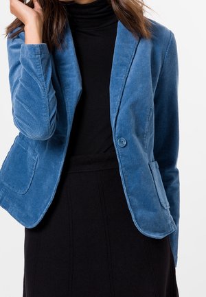 Femme portant un blazer en velours côtelé bleu par-dessus un col roulé noir et une jupe noire, la main gauche touchant ses cheveux.