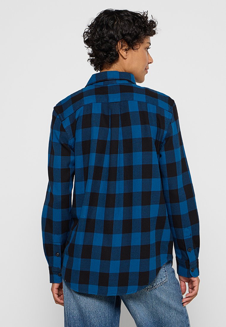 Persona con capelli corti e ricci girata di spalle, indossa una camicia a maniche lunghe a scacchi blu e nera e jeans blu.