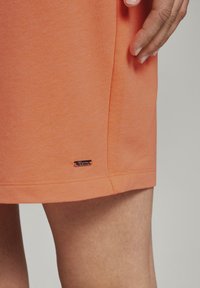 Falda de algodón naranja, hasta la rodilla, con textura suave. Presenta una pequeña etiqueta metálica cerca del dobladillo y detalles de costura.