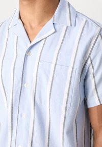 Gros plan d'une personne portant une chemise bleu clair à manches courtes avec des rayures verticales blanches et noires et une poche poitrine.