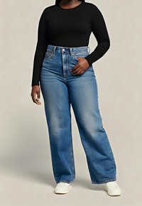 Femme portant un haut noir à manches longues, un jean bleu taille haute à jambes larges et des baskets blanches, debout devant un fond neutre.