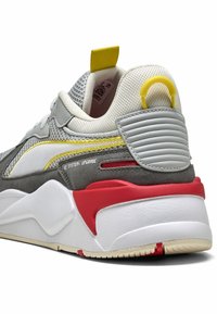 Szürke és fehér sneaker sárga részletekkel, texturált hálós felsőrésszel, párnázott sarkal és piros-fehér talppal, amelyen hangsúlyos hullámmintázat található.