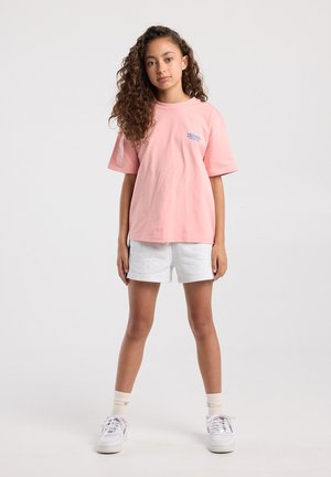 Jong meisje met krullend haar, staand, draagt een oversized roze T-shirt, lichtgrijze korte broek, witte sokken en witte sneakers.