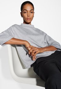 Pullover argentato con texture, colletto alto e maniche corte, abbinato a pantaloni neri. Modella seduta su una sedia moderna, braccia incrociate.