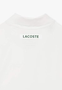 Hvid polo shirt med klassisk krave, blødt stof og en fremtrædende grøn "LACOSTE"-logo på nakken.