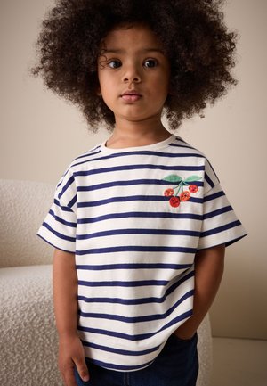 Fiatal gyermek göndör hajjal, fehér és tengerészkék csíkos pólót visel, mellkasán hímzett piros cseresznyékkel, beltéren áll.