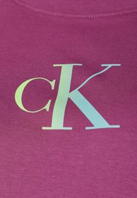 Fialová tkanina s hladkým povrchem, která má vícerozměrné logo CK s lesklým povrchem. Logo je výrazně umístěno uprostřed a má velké rozměry.