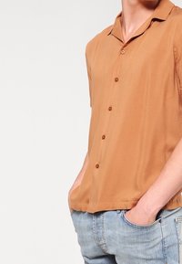 Kortärmad, ljusbrun skjorta med klassisk krage, fem knappar och avslappnad passform. Materialet verkar slätt med en subtil lyster.