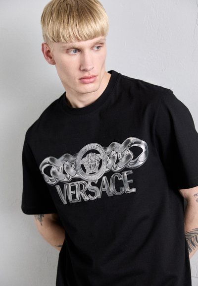 T-shirt en coton noir avec un graphique argenté de chaînes et le logo Versace sur la poitrine. Col rond et manches courtes.