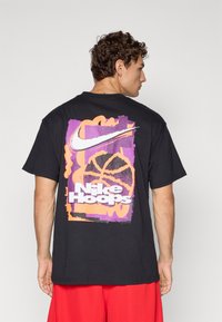 Homme portant un t-shirt noir Nike avec un graphique de basketball violet et orange et le texte "Nike Hoops", associé à un short rouge, tourné de côté.