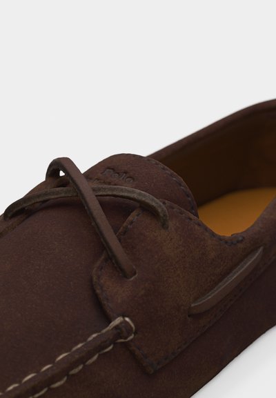 Καφέ μοκασίνι σουέτ με δερμάτινα κορδόνια, λεπτομερείς ραφές και ταμπά εσωτερική επένδυση.